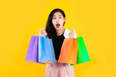 Young asian woman long hair carrying green, blue orange and purple shopping paper bag posing wow and excited on yellow background.