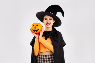 Cadılar Bayramı teması, siyah cadılar bayramı kostümü giymiş, elinde turuncu balkabağı kovası olan Asyalı genç ve güzel bir kadın izole edilmiş beyaz bir zemin oluşturuyor..