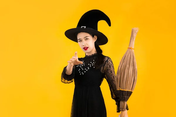 ハロウィンテーマ 若いです可愛いですアジアの女性で黒のドレス魔女衣装を身に着けています黒魔女帽子保持ほうきを持っている黄色の色の背景にポーズ — ストック写真