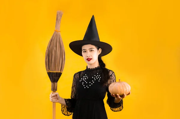 ハロウィンテーマ 若いアジア人女性で黒のドレス魔女衣装を身に着けている黒魔女帽子保持ほうきとカボチャのポーズ黄色の色の背景 — ストック写真