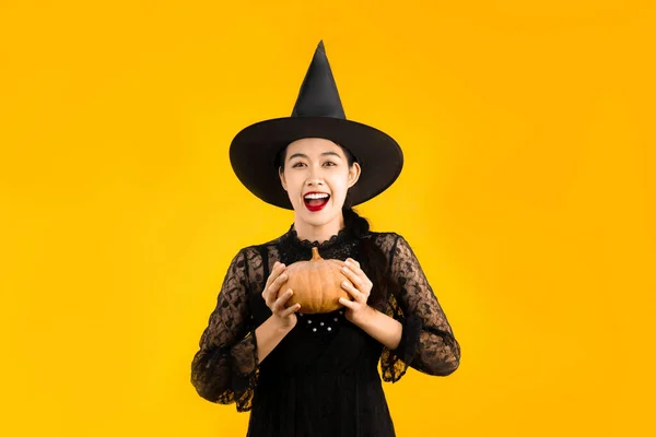 ハロウィンテーマ 幸せな若いアジアの女性で魔女衣装黒ドレス帽子を保持カボチャを身に着けている黄色の色の背景 — ストック写真