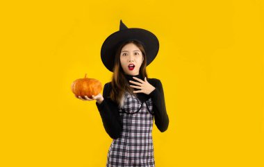 Halloween theme, young asian woman in black dress wearing witch hat holding orange pumpkin posing surprised and excited on yellow backgroud.	