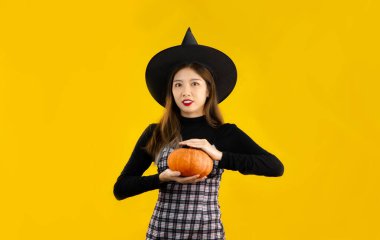 Cadılar Bayramı teması, siyah elbiseli genç Asyalı kadın. Cadı şapkası takıyor. Elinde turuncu balkabağı var. Sarı arka planda poz veriyor..