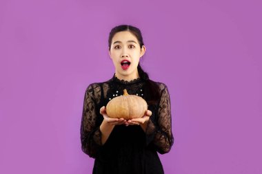 Halloween theme, young asian woman in black dress holding pumpkin on purple color background.	