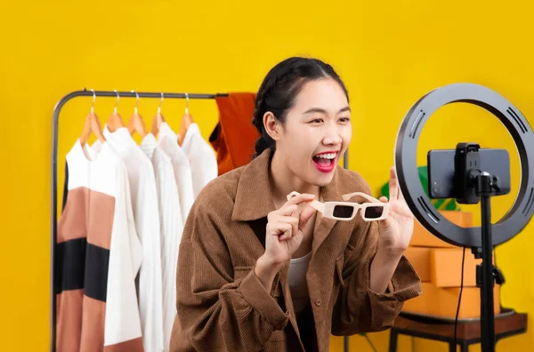 Live streaming concept, young asian blogger woman showing sunglasses in front of smartphone camera while recording vlog video live chat and selling on her online shop at home.