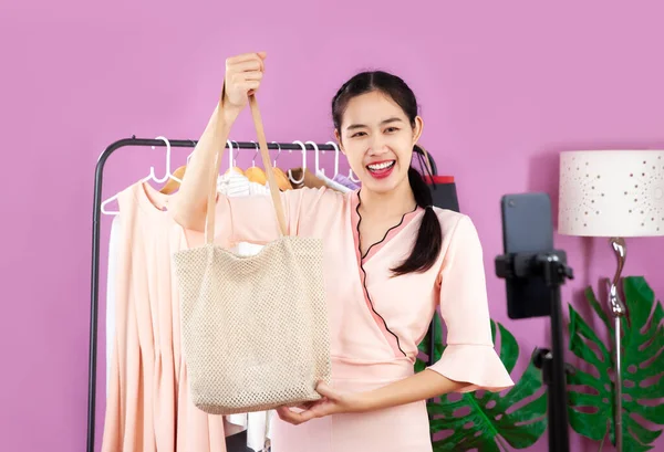 Live streaming concept, young asian blogger woman showing bag in front of smartphone camera while recording vlog video and live chat and selling at her online shop.
