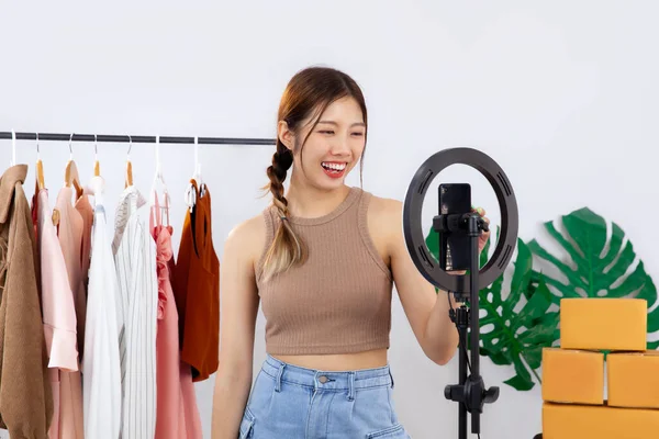 Young asian blogger woman live streaming in front of smartphone camera while recording vlog video and selling clothes on her online shop. Live streaming concept.