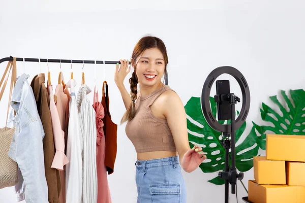 Young asian woman live streaming and chatting for online selling in front of smartphone camera while recording vlog video at home.