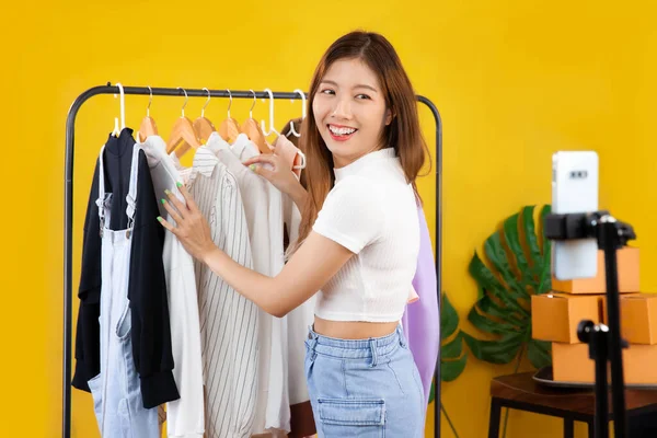 Young asian woman live streaming showing clothes in front of smartphone camera chatting and selling online shop yellow color background.