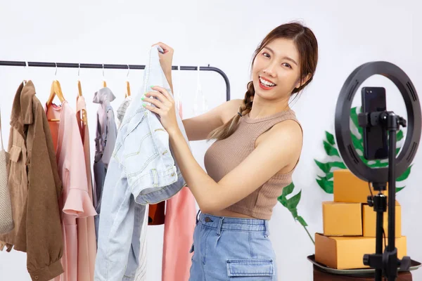 Live streaming concept, young asian blogger woman showing clothes in front of smartphone camera while recording vlog video live chat and selling at her online shop.