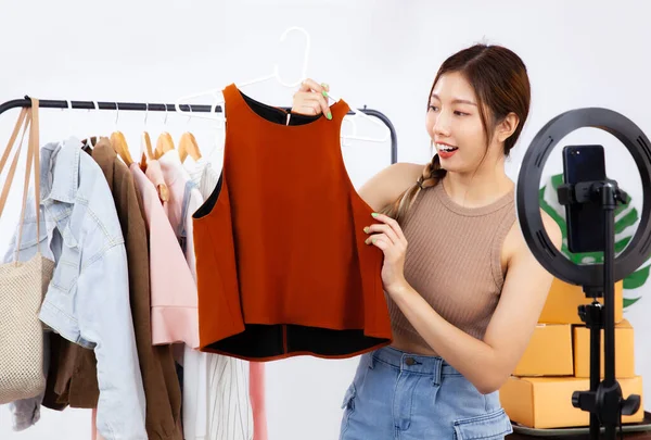 Live streaming concept, young asian blogger woman showing clothes in front of smartphone camera while recording vlog video and live chat and selling at her online shop.