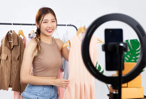 Live streaming, young pretty asian woman blogger showing clothes in front of smartphone camera while recording vlog video and live selling at her online shop.