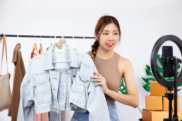 Young prettyy asian woman blogger showing clothes in front of smartphone camera while recording vlog video and live streaming at her shop. SME or freelance small business online and delivery concept.