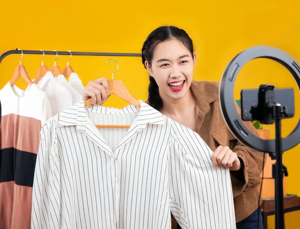Young asian woman blogger showing clothes in front of smartphone camera while recording vlog video and live streaming at her shop. Freelance or entrepreneur business online and delivery concept.