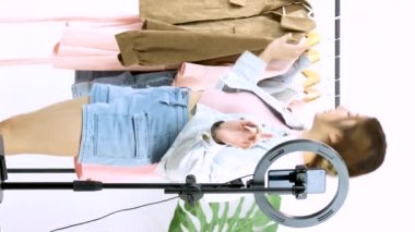 Canlı yayın, dikey video 4K. Genç Asyalı güzel kadın moda vlogcusu sohbet ediyor ve akıllı telefondan kıyafet satıyor. Evdeki pembe renkli duvar..