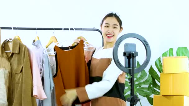 若いアジアの女性ファッションVloggerは 自宅で販売作業スマートフォンのカメラレコードビデオライブストリーミングオンラインショップに服を示しています ライブストリーミングビデオ — ストック動画
