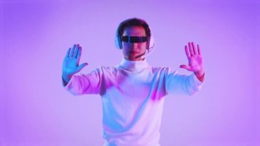 4K video Asyalı beyaz uzun kollu tişörtlü, VR gözlüklü, kulaklık takan, parmak gösteren ve mor arkaplana dokunan bir adam. Fütürist yaşamın metaverse konsepti.