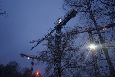 Akşam gökyüzünün arka planına karşı çalışan vinçler. Vinç genellikle kaldırma halatı, tel halatlar, zincirler ve demetler ile donatılmış bir makinedir, hem kaldırma hem de alttaki malzemeleri yatay olarak hareket ettirmek için kullanılabilir. Berlin