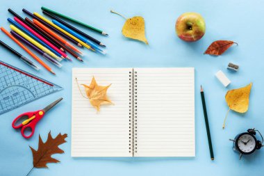 Boş not defteri ve okul malzemeleri, çalar saat, elma ve sarı sonbahar yaprakları mavi arka planda. Okul konseptine geri dönelim. Üst görünüm, düz görünüm, maket.