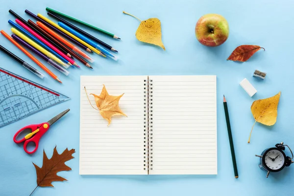 Boş not defteri ve okul malzemeleri, çalar saat, elma ve sarı sonbahar yaprakları mavi arka planda. Okul konseptine geri dönelim. Üst görünüm, düz görünüm, maket.