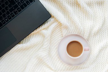 Beyaz örgü battaniyede bir fincan kahve ve bir defter bilgisayarı. Evden çalışmak, serbest meslek anlayışı. Üst manzara, düz uzanma