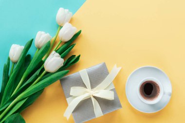 Beyaz laleler, hediye kutusu ve iki tonlu mavi ve sarı arka planda kahve fincanı. Bahar tatili, doğum günü, anneler günü konsepti. Üst görünüm, düz görünüm, kopyalama alanı.