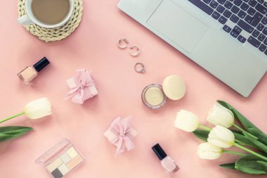 Laptop bilgisayar, kahve fincanı, hediye kutuları, kozmetik ürünler, aksesuarlar ve pembe arka planda çiçekler. Bahar tatilleri, online alışveriş konsepti. Üst manzara, düz uzanma.