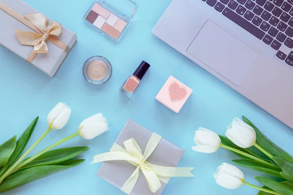Laptop, hediye kutuları, makyaj malzemeleri ve mavi arka planda lale çiçekleri. Bahar tatilleri, online alışveriş konsepti. Üst manzara, düz uzanma.