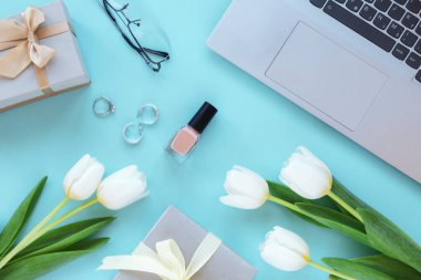 Laptop, bardaklar, hediye kutuları, makyaj malzemeleri ve mavi arka planda beyaz lale çiçekleri olan aksesuarlar. Bahar tatilleri, online alışveriş konsepti. Üst manzara, düz uzanma.