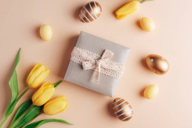 Paskalya bayramı geçmişi. Kutuda hediyeler, renkli Paskalya yumurtaları ve bej arka planda sarı laleler. Üst manzara, düz uzanma.