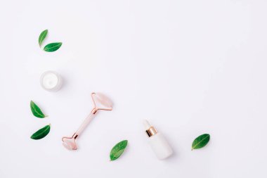 Kavanozda krema, serum şişesi ve beyaz arka planda yeşil yapraklı kristal gül kuvars yüz silindiri. Düz yat, boşluğu kopyala.