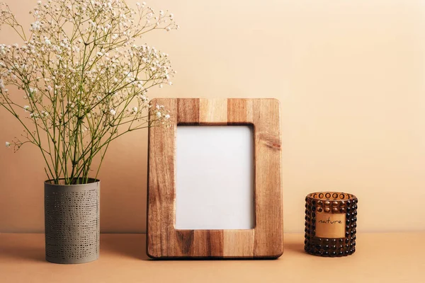 Boş resim çerçevesi, aroma mumu ve bej masadaki vazoda çingene çiçekleri. Ön görünüm, modelle