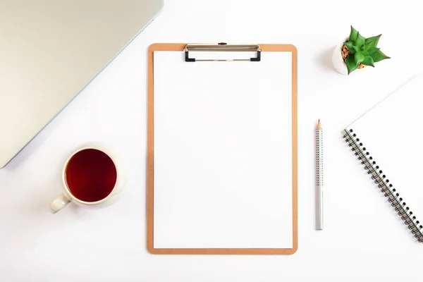 White desktop with clipboard, cup of tea, pencil, notebook and succulent plant. Flat lay, top view, mockup.