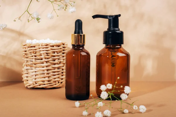 Kozmetik yüz temizleyici ve serum kahverengi cam şişeler sepette kulak çubukları ve açık bej arka planda çingene beyaz çiçekler. Doğal kozmetik, spa ve sağlık konsepti. Kapat..