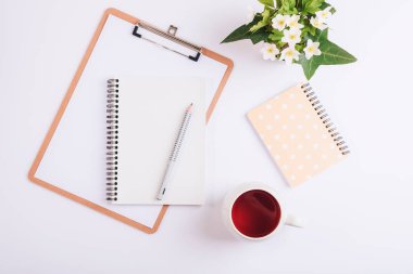 Kağıt panosu, çay fincanı, kalem, defter ve bir buket bahar çiçeği olan beyaz bir masaüstü. Düz yatış, üst görünüm, model