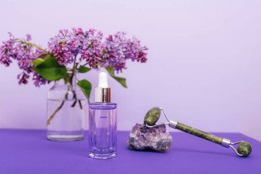 Kozmetik serum şişesi, yeşim yüz silindiri ve mor arka planda leylak çiçekleri. Doğal kozmetik ürünleri, cilt bakımı konsepti.