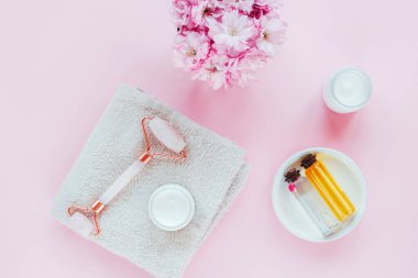 Sakura çiçekli doğal organik kaplıca seti. Yüz silindiri, kozmetik kremi, pembe arka planda havlu ve yağ. Cilt bakımı, güzellik tedavi konsepti. Üst manzara, düz uzanma.