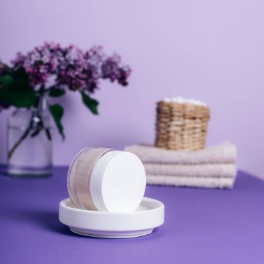 Yüz kremi kavanozu, havlular ve mor arka planda leylak çiçekleri. Seçici odak.