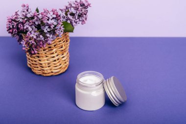 Krema kavanozu ve mor arka plandaki sepetteki leylak çiçekleri. Doğal kozmetik konsepti. Kapat..