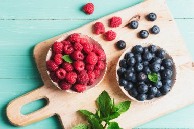 Mavi ahşap masadaki kesme tahtasının üstünde taze yaban mersini ve ahududu kaseleri. Sağlıklı beslenme, vejetaryen yemekleri..