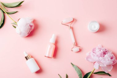 Kozmetik yağı, serum, yüz silindiri, krema kavanozu ve pembe arka planda şakayık çiçekleri olan damlalar. Üst manzara, düz uzanma.