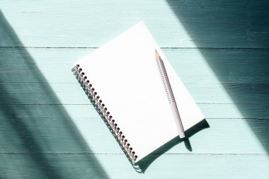 Boş çarşaf ve turkuaz mavi ahşap masa üzerindeki kalemle güneş ışığı altında not defteri. Üst görünüm, düz görünüm, model.