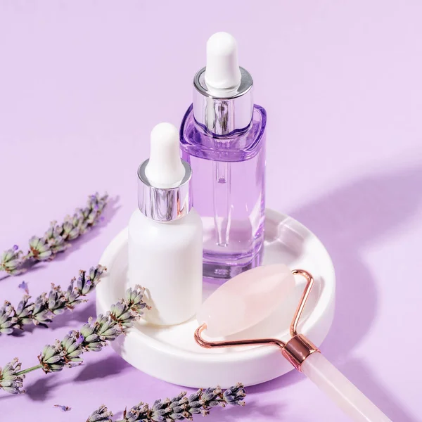 Skincare products, serum and cosmetic oil, rose quartz crystal facial roller and lavender blossom on light purple background. Aromatherapy and natural cosmetics concept. Closeup.