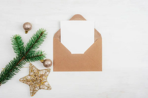 Open brown envelope with blank white card and Christmas decorations on white table background. Top view, flat lay, mockup.