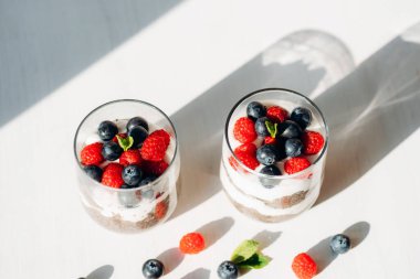 Beyaz masada taze ahududu ve yaban mersinli iki bardak chia pudingi. Uzun gölgeler, üst manzara.