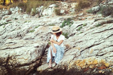 Girl in a straw hat sits on a rock by the sea on a sunny summer day.