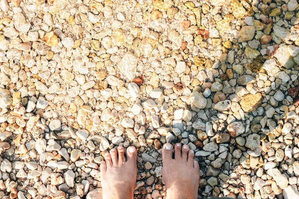 Feet on a pebbly beach at the water\'s edge.