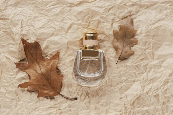 Perfume bottle and autumn leaves on beige craft paper background. Top view, flat lay.