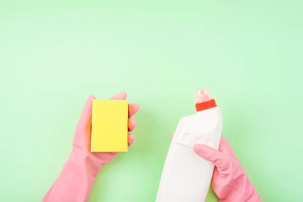 Cleaners hand in pink rubber protective glove holding bottle with detergent and yellow sponge on green background. Top view.