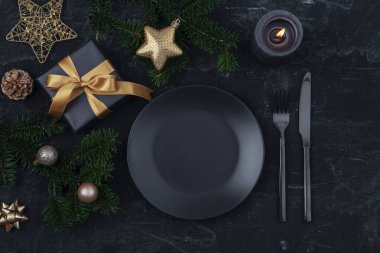 Şık masa dekorasyonu, hediye kutusu, köknar dalları, siyah tabaklı yıldız ve toplar, çatal bıçak takımı ve mum. Noel konsepti. Üst manzara, düz uzanma.